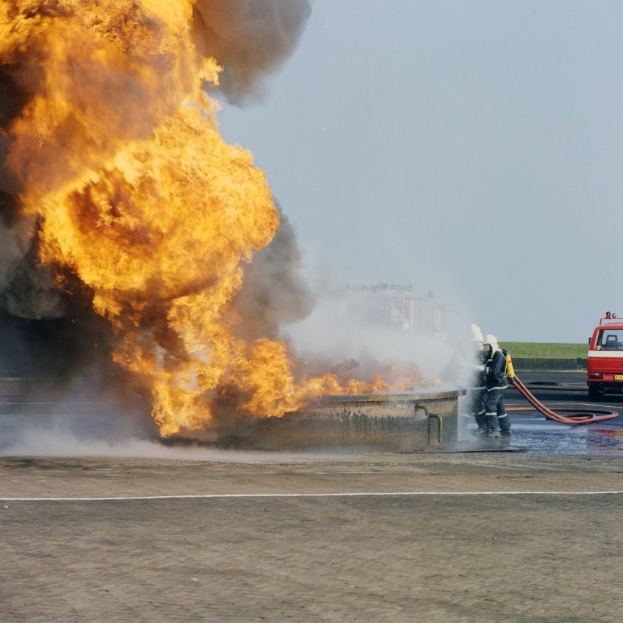 Feuerwehrl├Âschfahrzeug von Flammen umgeben auf der Seite einer Stra├če mit zwei Helmen tragenden Individuen, die Rohre halten, einem Fahrzeug im Hintergrund und dem Himmel.