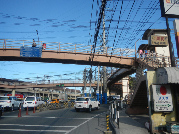 Eine belebte Straße mit einer Brücke darüber, mit Verkehrskegeln, Pfählen, Schildern, Passanten und Strommasten mit Drähten, vor Gebäuden und einem klaren blauen Himmel.
