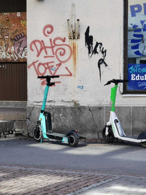 Zwei Elektro-Scooter nebeneinander auf einer Straße geparkt mit einer Graffiti-bedeckten Wand und einem Plakat im Hintergrund.