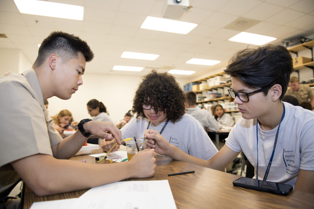 Three students at a table in a classroom working on a project with papers, pens, a mobile phone, and other objects on the table, and racks with items in the background under ceiling lights and a projector.
