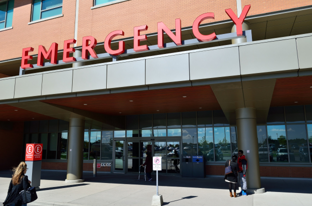 Eine Gruppe von Menschen, die vor einem Notfallgebäude mit Glaswänden und Säulen, einem Namensschild, Schildern auf Pfählen und einem Mülleimer gehen.