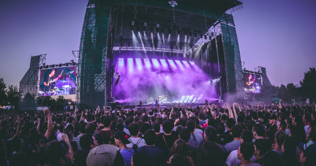 Eine große Menge Menschen auf einem Musikfestival, mit einer Bühne in der Mitte, auf der Musiker Instrumente spielen, beleuchtet von Lichtern und umgeben von Bildschirmen, mit einem Gebäude, Bäumen und einem klaren blauen Himmel im Hintergrund.