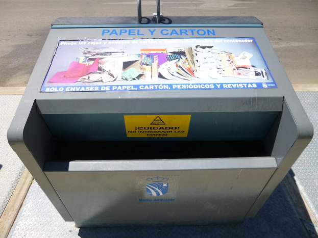 Metallmülltonne am Straßenrand mit einem "Papel y Carton"-Plakat, Fahrzeuge im Hintergrund sichtbar.