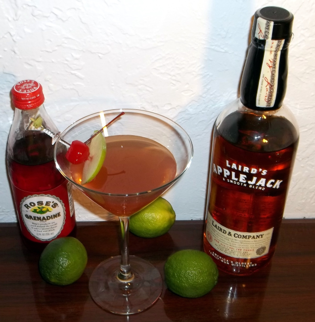 Martini in a coupe glass with cherry and lime garnish on a table next to a liquor bottle, against a wall background.