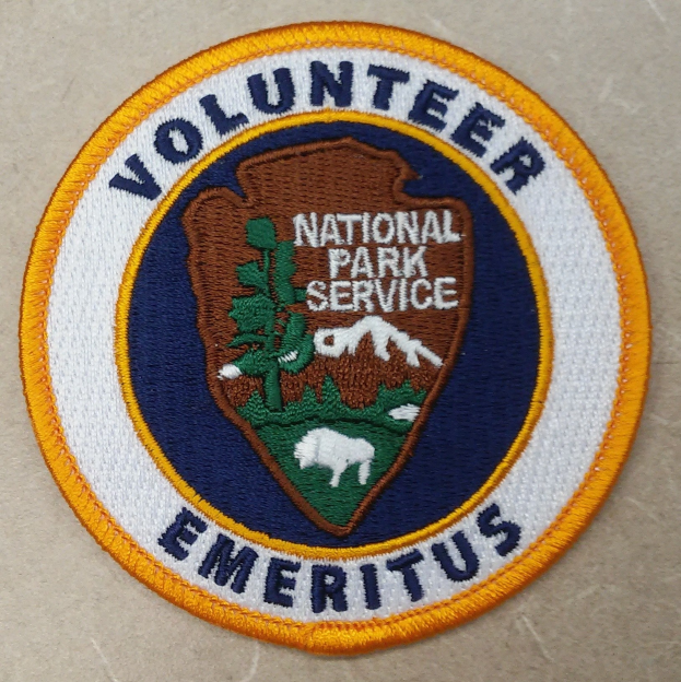 Runder Patch mit "Ehrenamtlicher Nationalparkdienst Emeritus" in fetter schwarzer Schrift auf weißem Hintergrund, eingerahmt von einem blauen Rand und einem gelben Kreis.