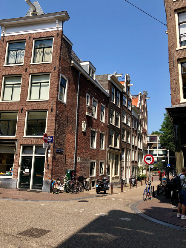 Eine Stra├če in Amsterdam, Niederlande, mit Geb├Ąuden auf beiden Seiten, Menschen, die gehen, geparkte Fahrr├Ąder, Schilder, B├Ąume und einen klaren blauen Himmel.