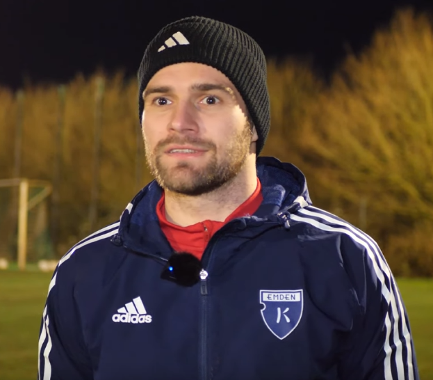 Ein Mann in einer blauen Jacke und Mütze steht auf einem Fußballfeld während eines Trainings, mit Bäumen und einem Tor im Hintergrund.