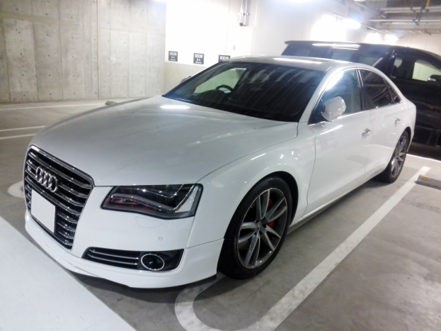 Weißer Audi S8 in einer Parkgarage neben einem schwarzen Auto geparkt, mit sichtbaren Garagenwänden, Deckenrohren und Lampen.
