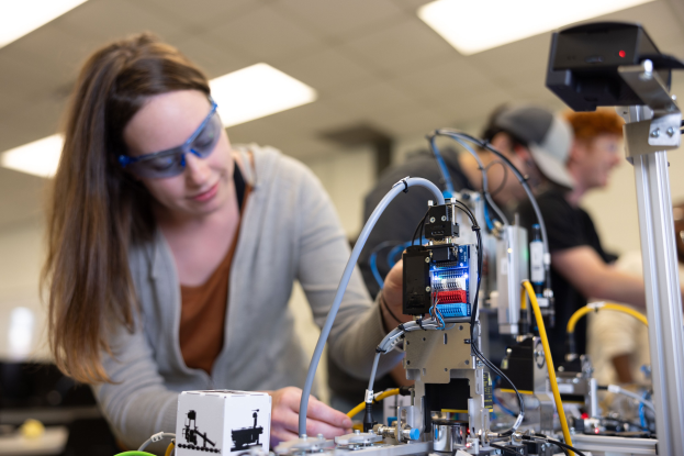 Eine Frau mit Schutzbrille arbeitet an einem Roboter in einem Labor, umgeben von anderen Personen und Gegenständen auf dem Tisch sowie Deckenbeleuchtung.