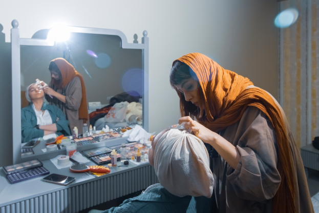 Eine Frau sitzt auf dem Boden vor einem Schminktisch mit Spiegel und hält dabei ein Objekt in der Hand; der Tisch ist voller Make-up-Bürsten, Flaschen, einem Handy, einem Taschenrechner und anderen Gegenständen, mit einer Lampe an der Wand und einem Vorhang auf der rechten Seite.