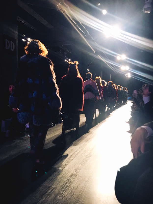 Eine Gruppe von Menschen läuft über den Laufsteg einer Fashion Show, beleuchtet von hellen Scheinwerfern, mit einem Publikum auf beiden Seiten.