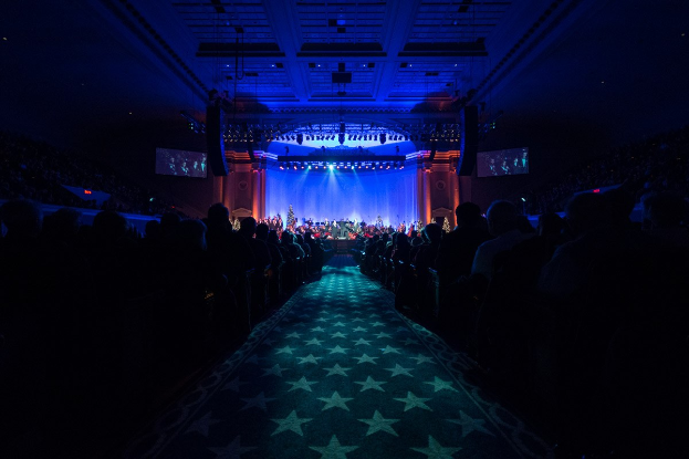 Ein großer Saal mit sitzenden Zuschauern, eine Bühne mit Musikern und Deckenbeleuchtung und -bildschirme, was auf einen Konzertsaal während der Nationalen Convention 2019 hindeutet.
