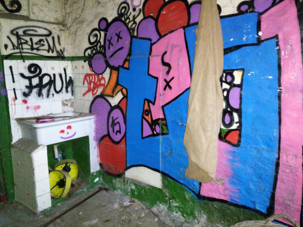 Ein Badezimmer mit Graffiti an den Wänden, ein Waschbecken in der Ecke, das mit einem Tuch bedeckt ist, und verschiedene Gegenstände, die darum herum verstreut sind.