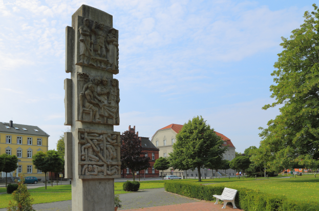 Ein steinernes Denkmal steht in einem Park mit grünem Gras, Pflanzen und Bäumen, mit einer Bank davor; Gebäude, Fahrzeuge und ein bewölkter Himmel sind im Hintergrund zu sehen.
