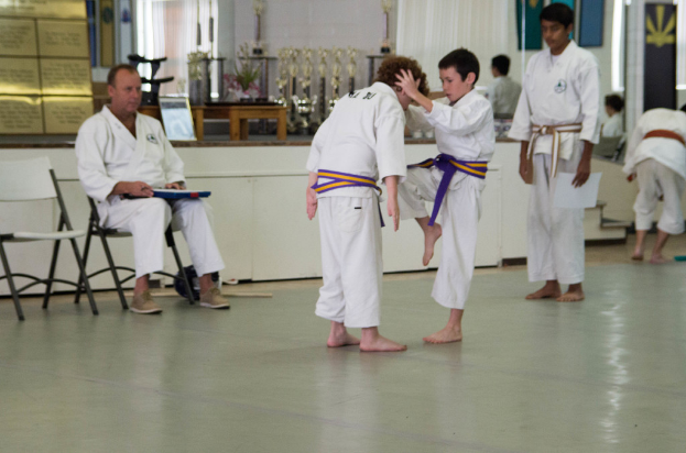 Zwei Kinder bei der Kung-Fu-Übung mit ihrem Meister, der links am Tisch sitzt und etwas prüft.